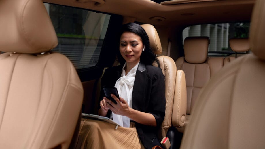 Woman sitting in the backseat of a car, looking at her smartphone and holding a tablet on her lap.