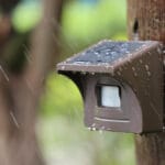 Close-up of eMACROS Pro 4 HS006W solar-powered outdoor sensor with water droplets on it, mounted on a wooden post in the rain.