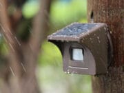Close-up of eMACROS Pro 4 HS006W solar-powered outdoor sensor with water droplets on it, mounted on a wooden post in the rain.