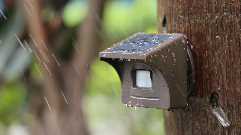 Close-up of eMACROS Pro 4 HS006W solar-powered outdoor sensor with water droplets on it, mounted on a wooden post in the rain.