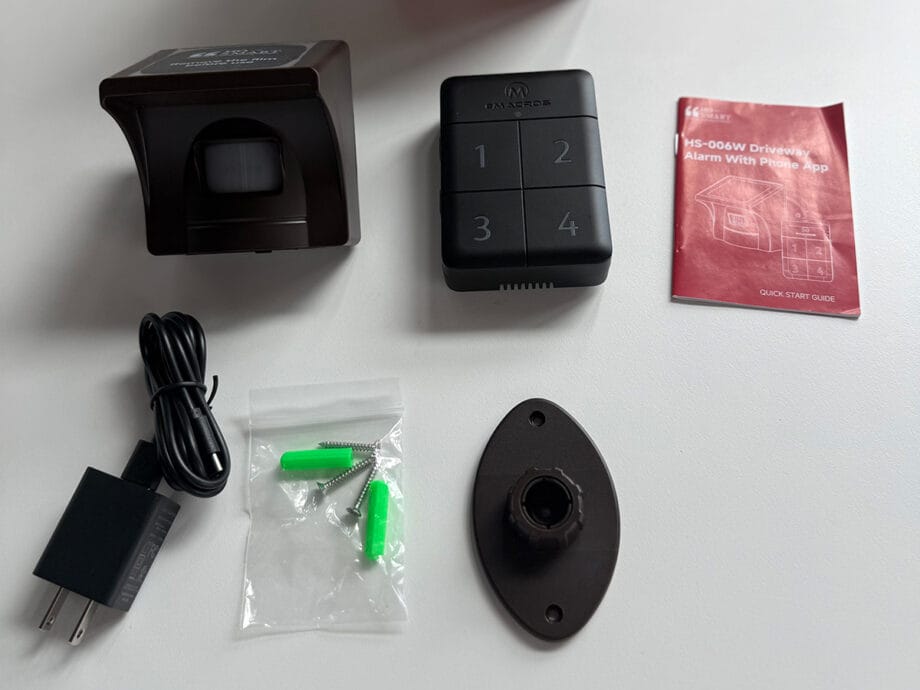 Components of eMACROS Pro 4 HS006W driveway alarm system laid out, including sensor, keypad, power adapter, screws, wall mount, and instruction manual.