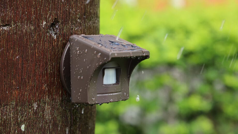 eMACROS Pro 4 HS006W outdoor motion sensor attached to a wooden post, with water droplets on the surface, in rainy weather.
