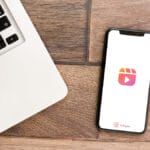 A smartphone displaying the Instagram Reels logo lies on a wooden surface next to a laptop keyboard.