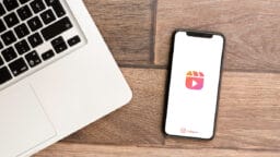 A smartphone displaying the Instagram Reels logo lies on a wooden surface next to a laptop keyboard.