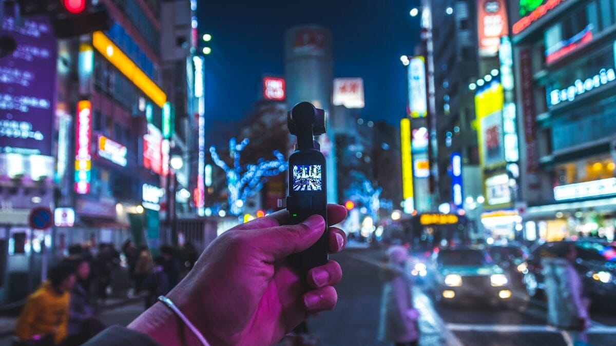 A hand holds a small camera, capturing a busy city street with bright neon lights and people crossing at night.