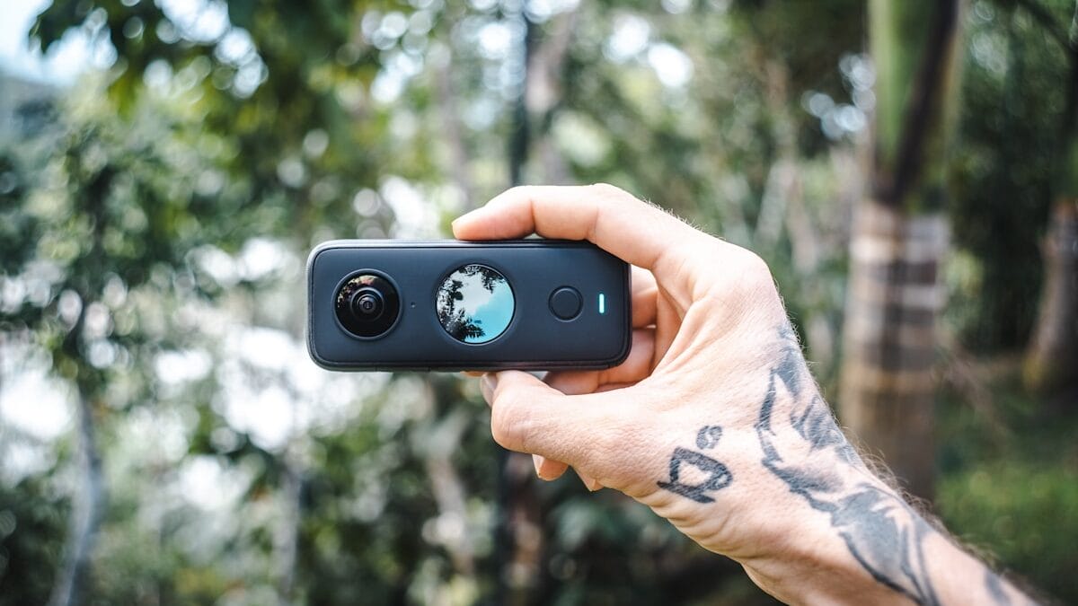 A tattooed hand holds a small 360-degree action camera outdoors, with trees and greenery blurred in the background.