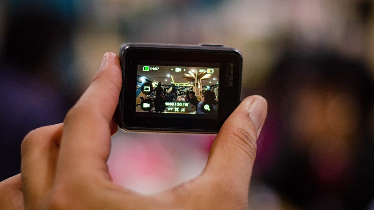 A hand holds a small camera displaying a viewfinder image of people gathered indoors under bright lights.