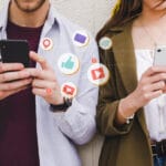 Two people use smartphones with social media icons such as likes, messages, and notifications illustrated between them.