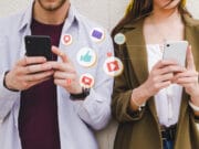 Two people use smartphones with social media icons such as likes, messages, and notifications illustrated between them.