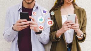 Two people use smartphones with social media icons such as likes, messages, and notifications illustrated between them.