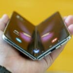 A hand holding a partially folded foldable smartphone with a visible hinge, against a yellow background.