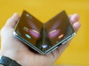 A hand holding a partially folded foldable smartphone with a visible hinge, against a yellow background.