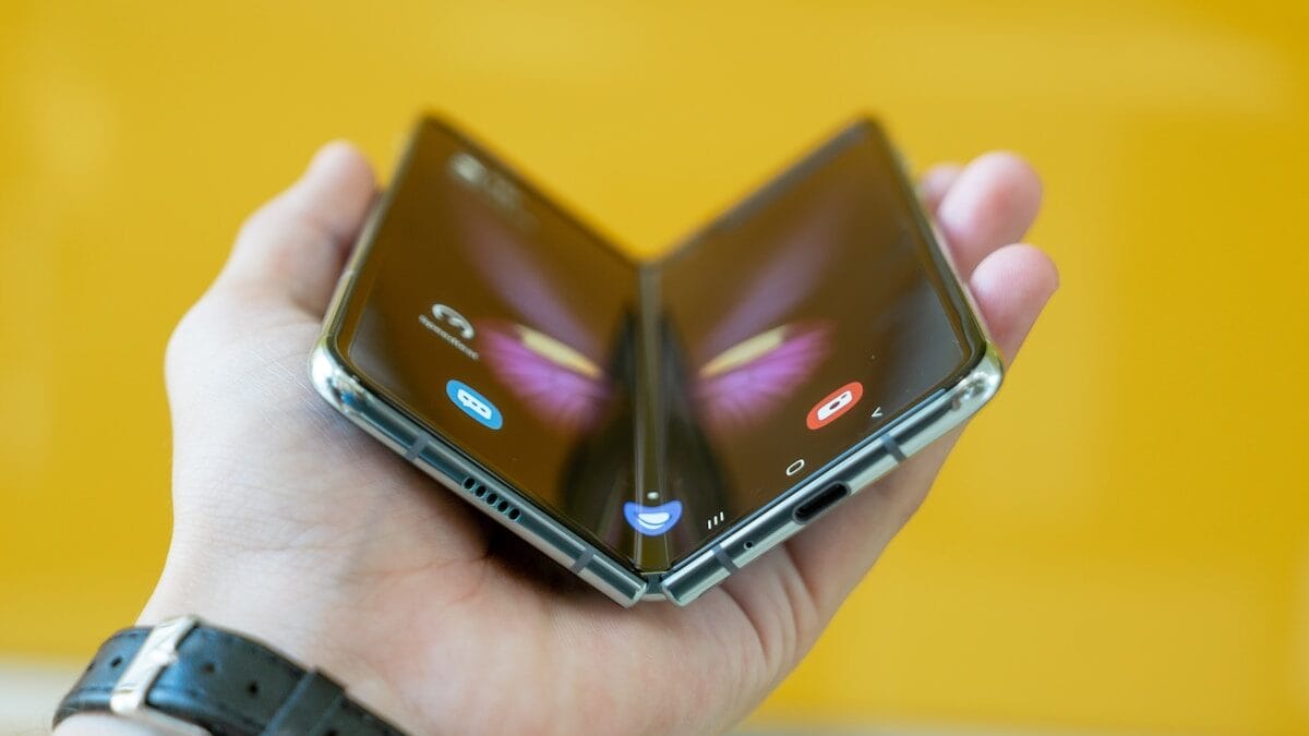 A hand holding a partially folded foldable smartphone with a visible hinge, against a yellow background.