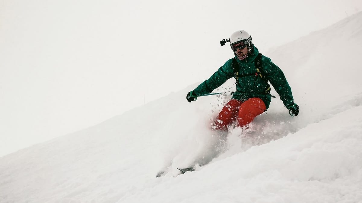 A person wearing a helmet and goggles skis downhill through deep snow, creating a spray of powder around them.