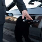 Person in a suit opening the rear door of a black car, with reflections visible on the car’s shiny surface.