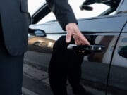 Person in a suit opening the rear door of a black car, with reflections visible on the car’s shiny surface.