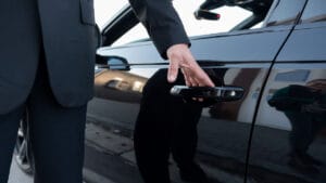 Person in a suit opening the rear door of a black car, with reflections visible on the car’s shiny surface.