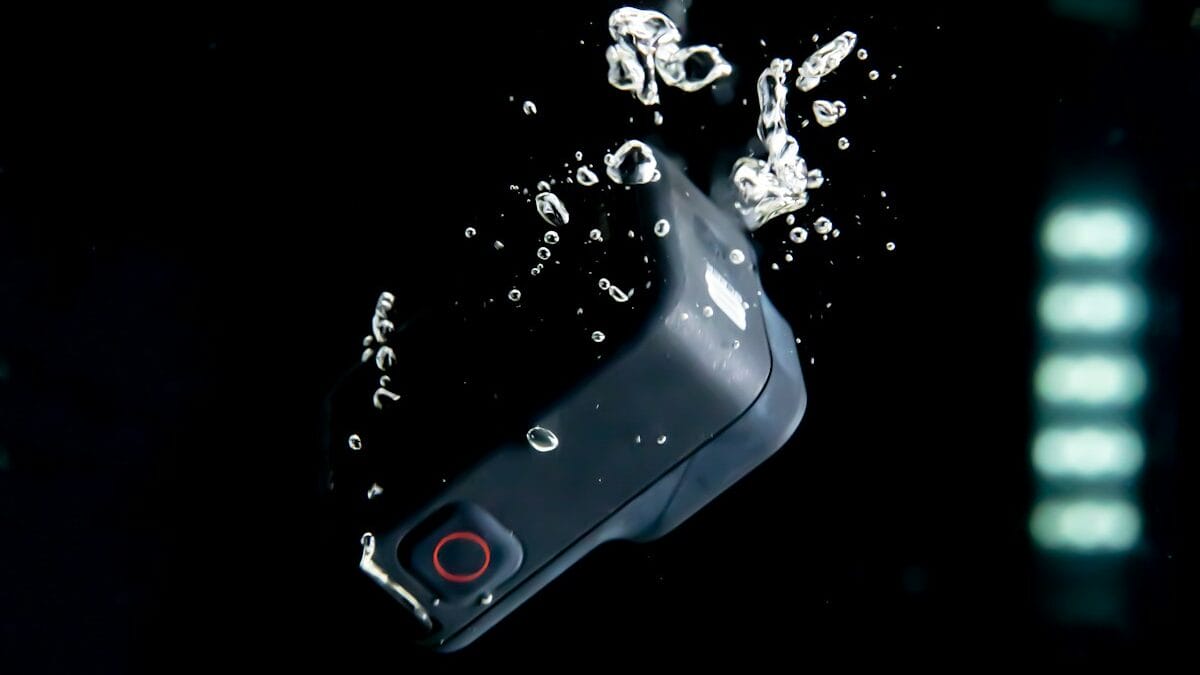 A small action camera is submerged underwater with bubbles rising around it, against a dark background.