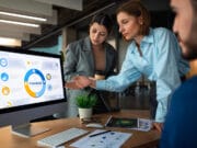 Three people in business attire discuss financial data displayed on a computer monitor in a modern office setting.