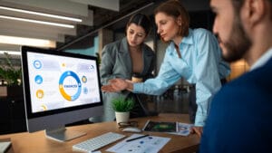 Three people in business attire discuss financial data displayed on a computer monitor in a modern office setting.