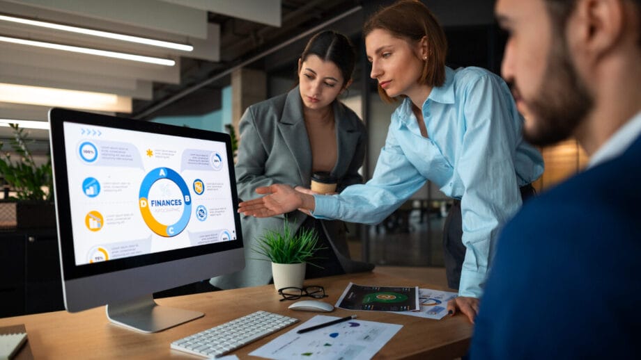 Three people in business attire discuss financial data displayed on a computer monitor in a modern office setting.