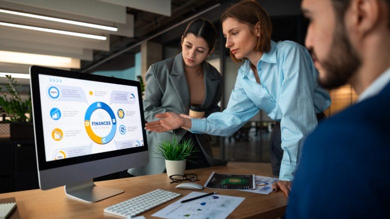 Three people in business attire discuss financial data displayed on a computer monitor in a modern office setting.