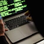 Person in dark clothing uses a smartphone and laptop with green code on the screen, suggesting computer programming or hacking.