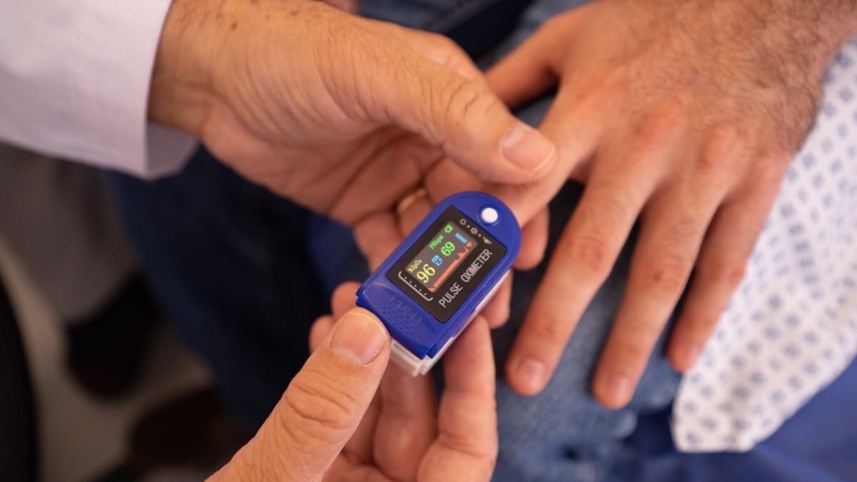 A person’s finger is placed in a pulse oximeter held by another person, displaying oxygen saturation and pulse rate.