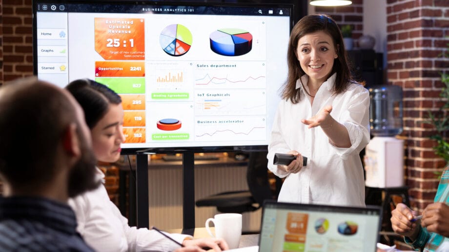 A woman presents business analytics charts on a screen to a group of colleagues in a modern office setting.