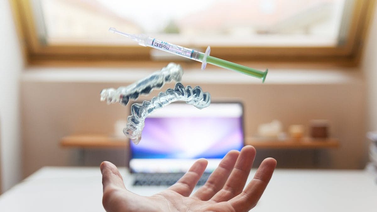 A hand tosses a syringe and two clear dental aligners in front of an open laptop on a desk.