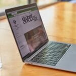 A glass of water sits next to an open laptop displaying a website called sweetlife on a wooden table by a window.