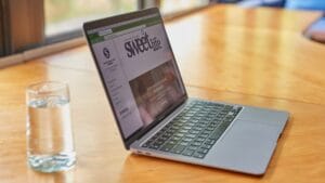 A glass of water sits next to an open laptop displaying a website called sweetlife on a wooden table by a window.