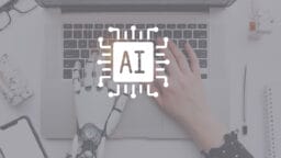 A human hand and a robotic hand type on a laptop keyboard, with an AI microchip icon overlay at the center.