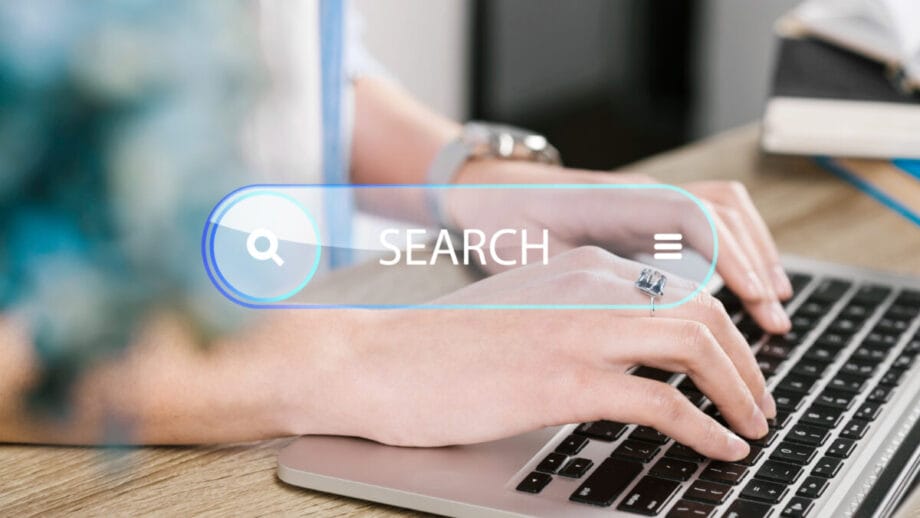 Hands typing on a laptop keyboard with a digital search bar overlay displaying the word SEARCH and a magnifying glass icon.