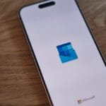 A smartphone on a wooden surface displays the Microsoft Outlook logo on its screen.