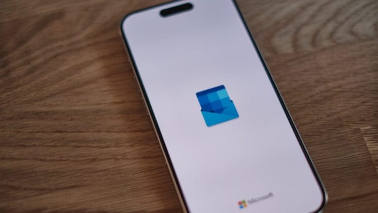 A smartphone on a wooden surface displays the Microsoft Outlook logo on its screen.