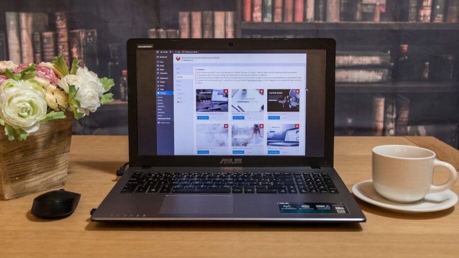 A laptop on a wooden table displays a website dashboard, with a mouse, potted flowers, and a cup of coffee nearby. Bookshelves in the background.