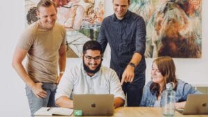 Four people gather around a table, smiling and working together on laptops in a bright office setting with artwork in the background.