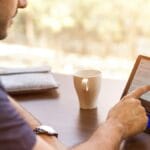 A person uses a touchscreen tablet at a desk with a notebook, coffee cup, and blurred outdoor background.