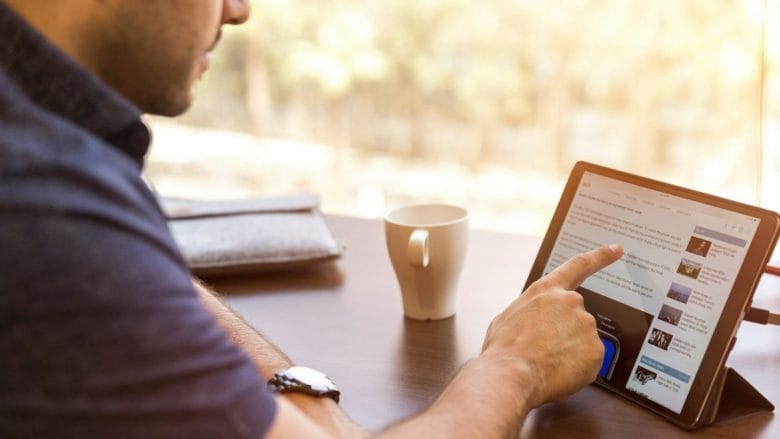 A person uses a touchscreen tablet at a desk with a notebook, coffee cup, and blurred outdoor background.