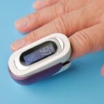 A hand with a fingertip pulse oximeter displaying oxygen saturation and pulse rate on a blue surface.