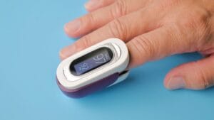 A hand with a fingertip pulse oximeter displaying oxygen saturation and pulse rate on a blue surface.