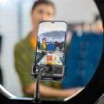 A person records themselves with a smartphone on a tripod, framed by a ring light, in what appears to be a retail or studio setting.