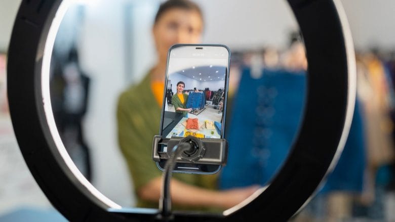A person records themselves with a smartphone on a tripod, framed by a ring light, in what appears to be a retail or studio setting.