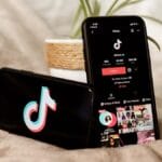 Two smartphones on a blanket; one displays the TikTok logo, the other shows a TikTok profile page, with a potted plant in the background.