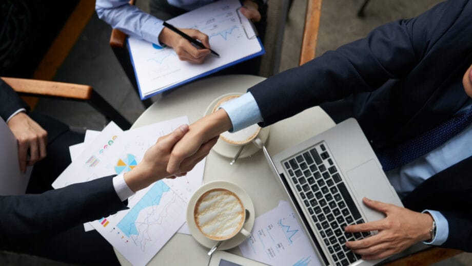 Two people in business attire shake hands over a table with documents, coffee cups, a laptop, and charts.
