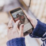 Person in a blue sweater holding a smartphone and viewing a photo of a woman on Instagram outdoors.