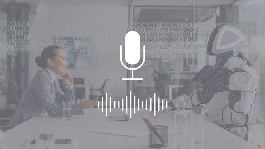 A woman and a robot sit across from each other at a table in an office, with a microphone and soundwave graphic overlay.