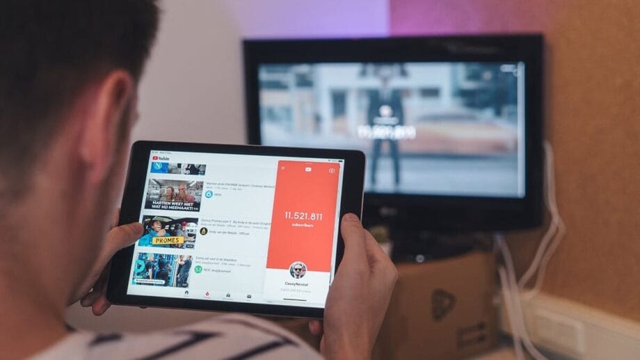 A person using a tablet to browse YouTube, with a TV screen displaying a blurred image in the background.