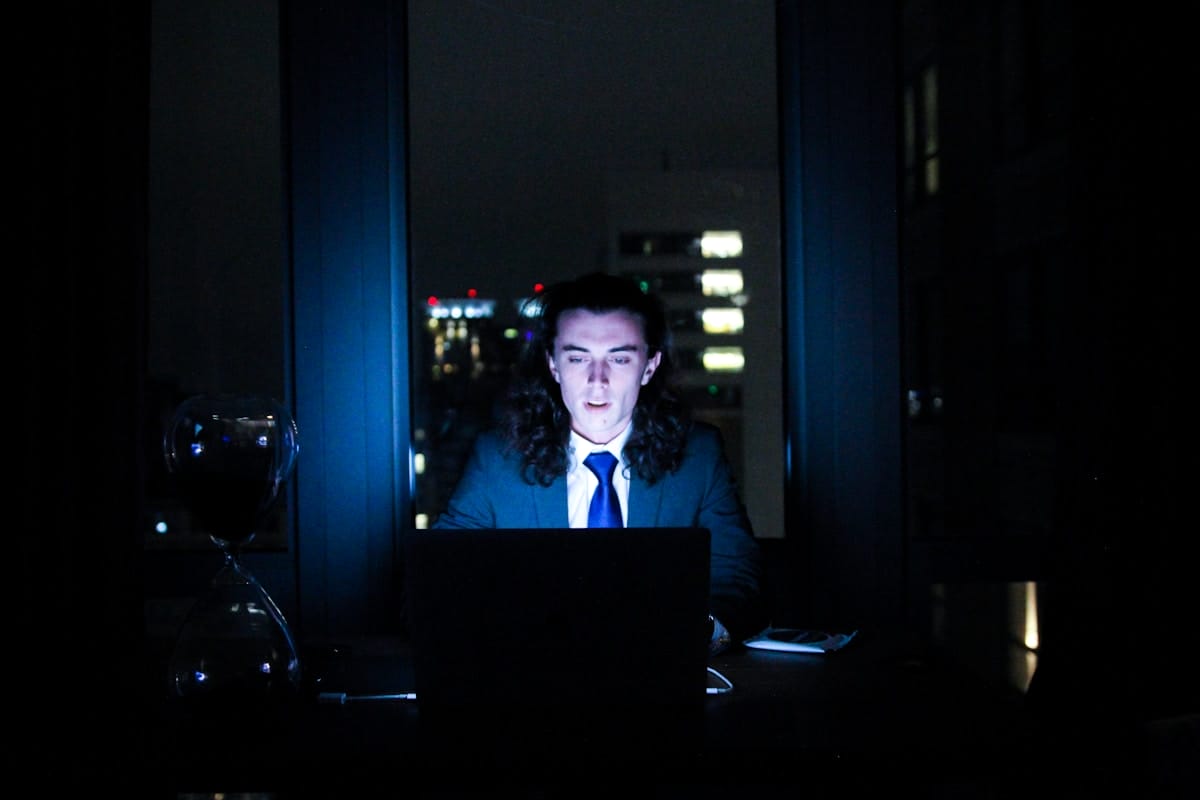A person in a suit works on a laptop at night in a dark room, illuminated by the screen, with an hourglass and phone nearby.